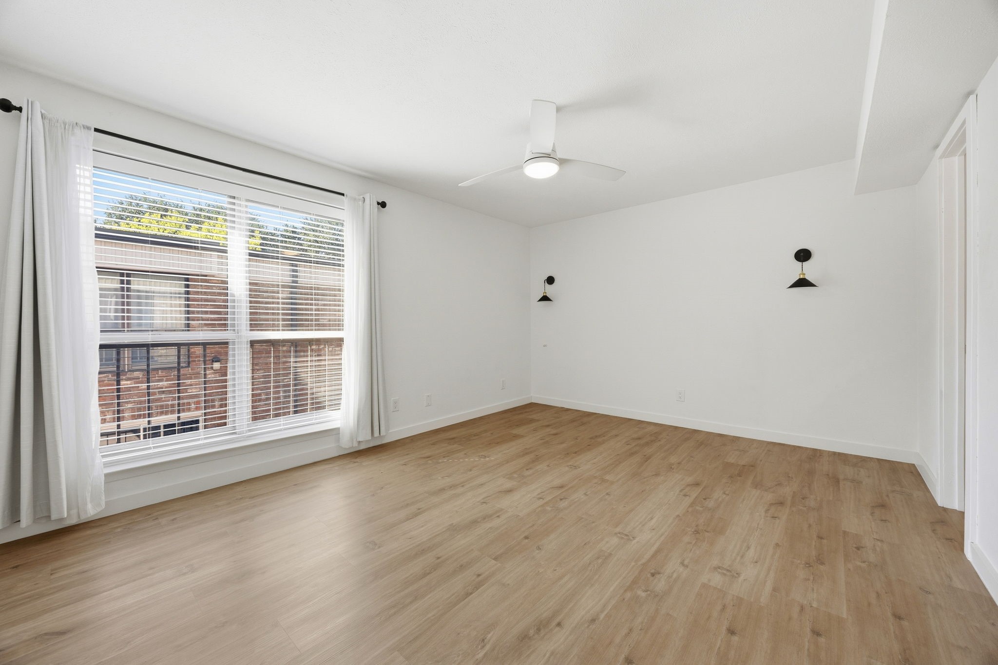 Vacant room with light wood floors, a large curtained window, wall sconces, and a ceiling fan.