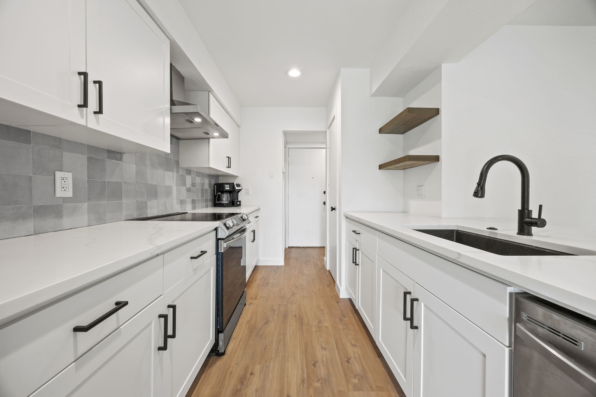 Galley kitchen with white cabinets, gray tile backsplash, black sink and faucet, and wood-look floors.