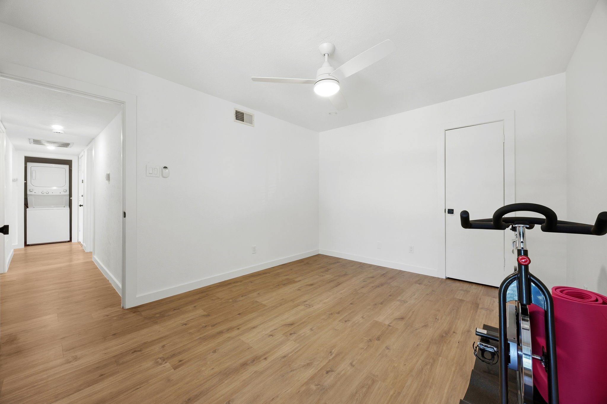Room with wood-look flooring and ceiling fan, exercise bike and yoga mat, and laundry appliances in the hallway.