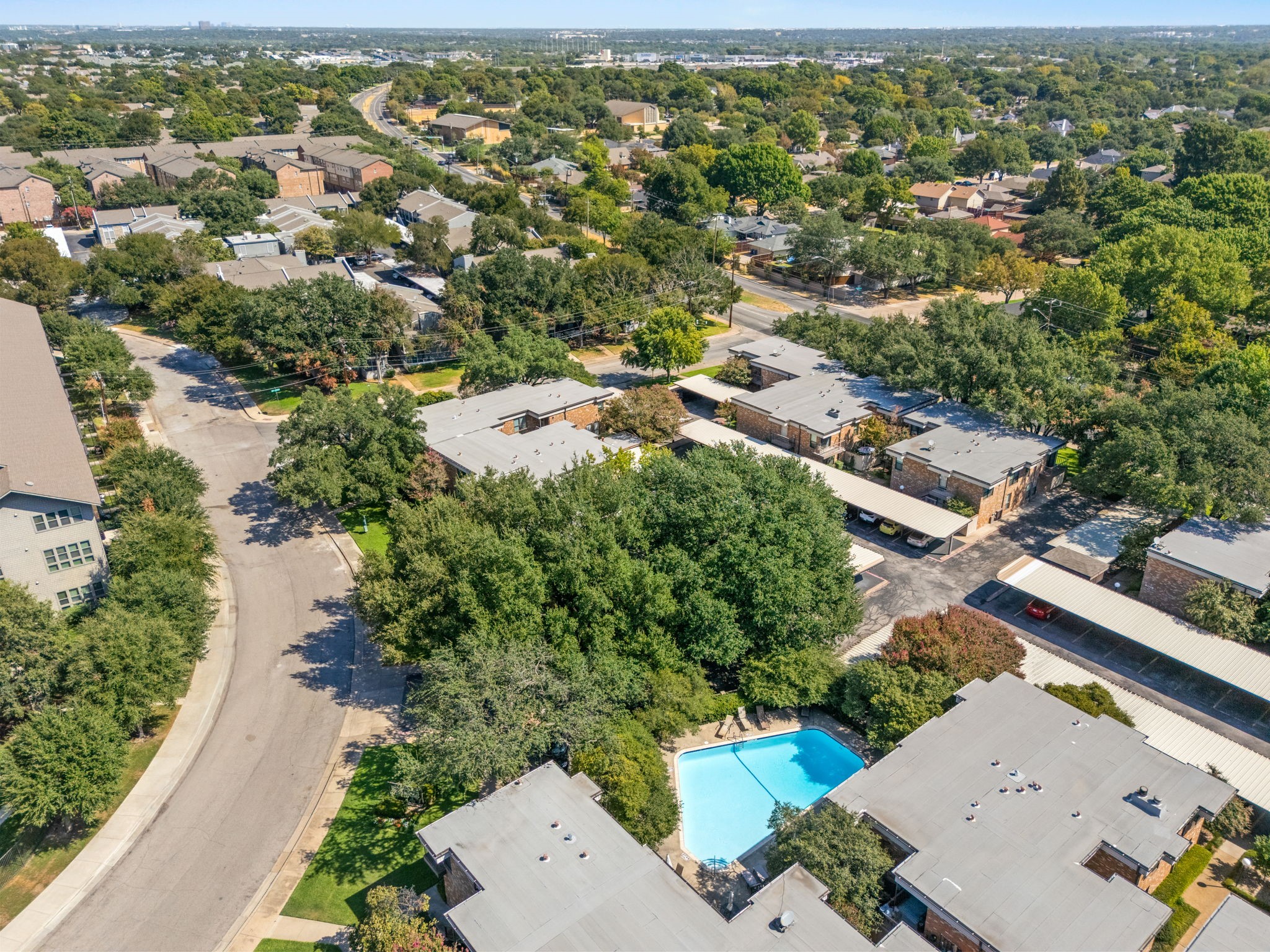 Aerial view of a tree-shaded condominium complex with a swimming pool and covered parking.