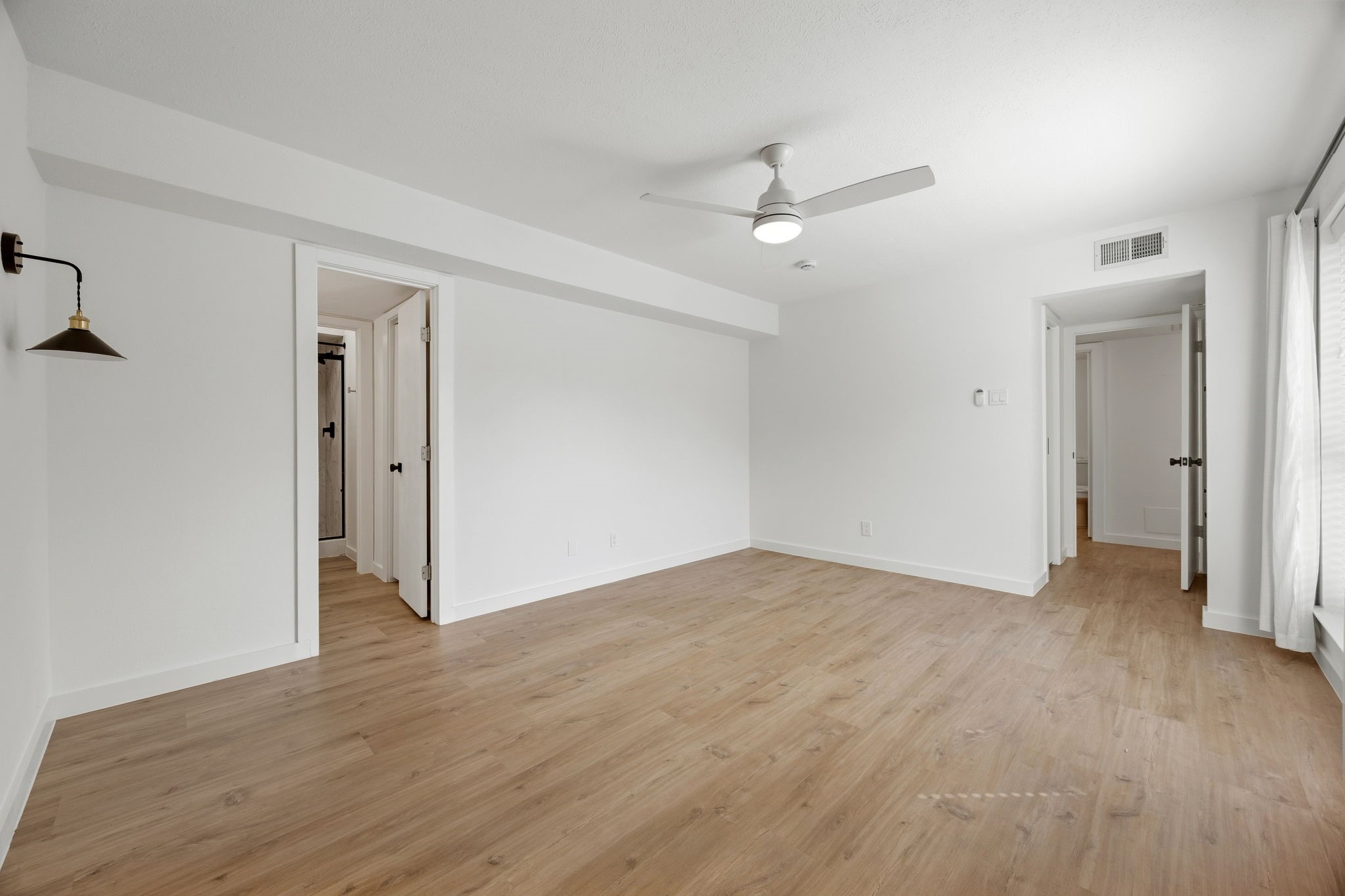 Empty bright room with light wood floors, a ceiling fan, and tall curtained windows.