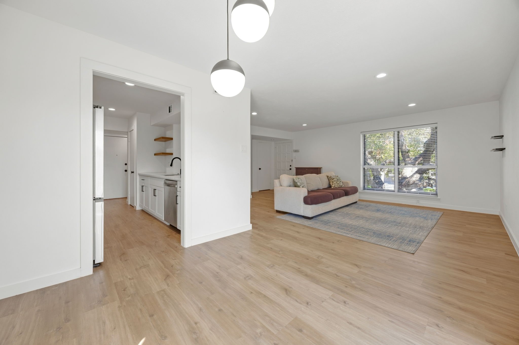 Open-plan living room with a sofa, large window, light wood floors, and kitchen beyond.