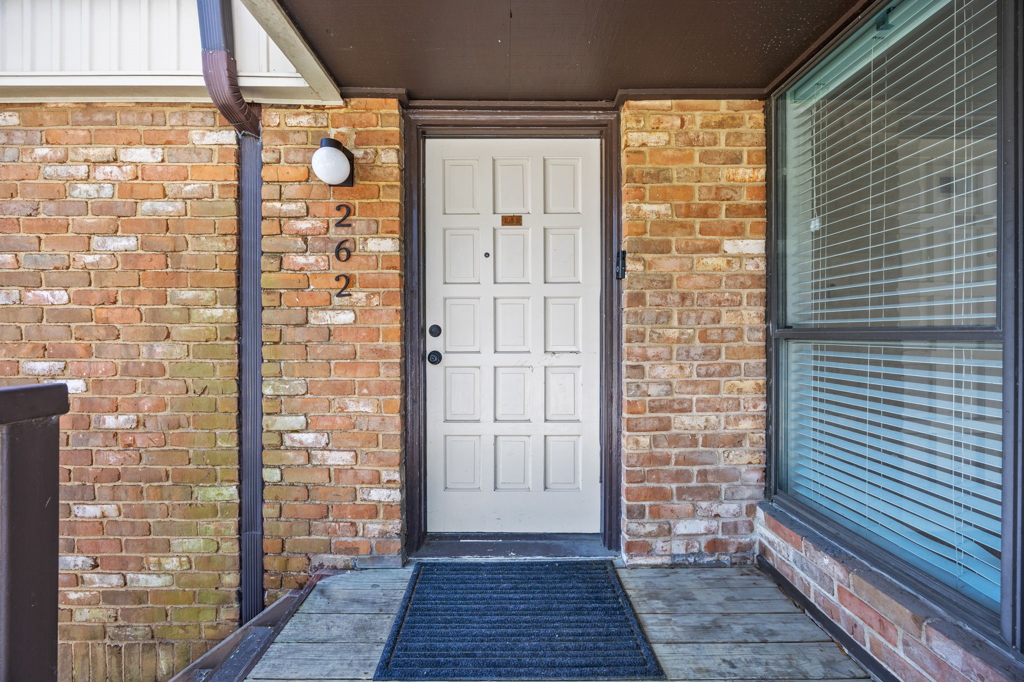 Brick residential entrance with a white paneled door marked 262 and a large window with blinds.