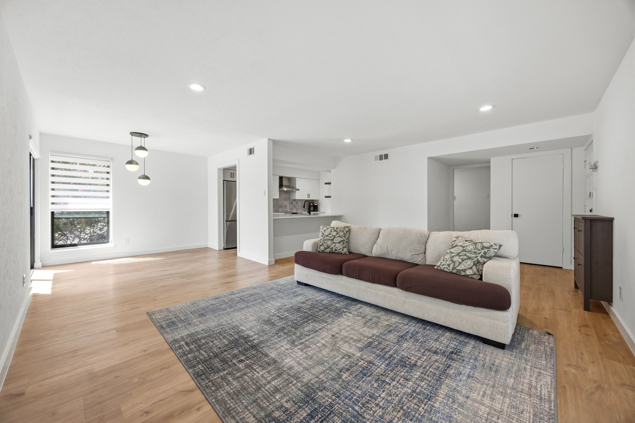 Open-plan living room with a cream sofa, patterned rug, light wood floors, and kitchen.