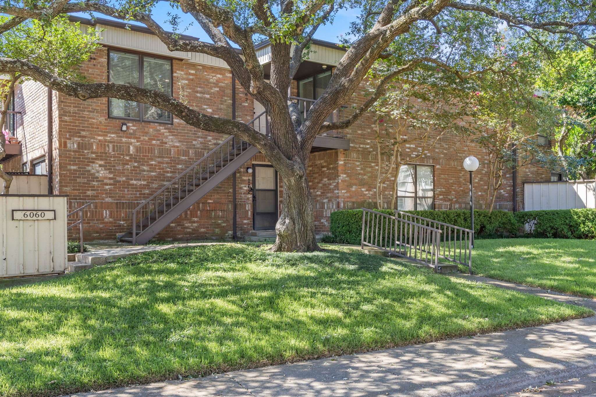 Two-story brick condominium with exterior stairs, a large shade tree, and a lawn-side access ramp.