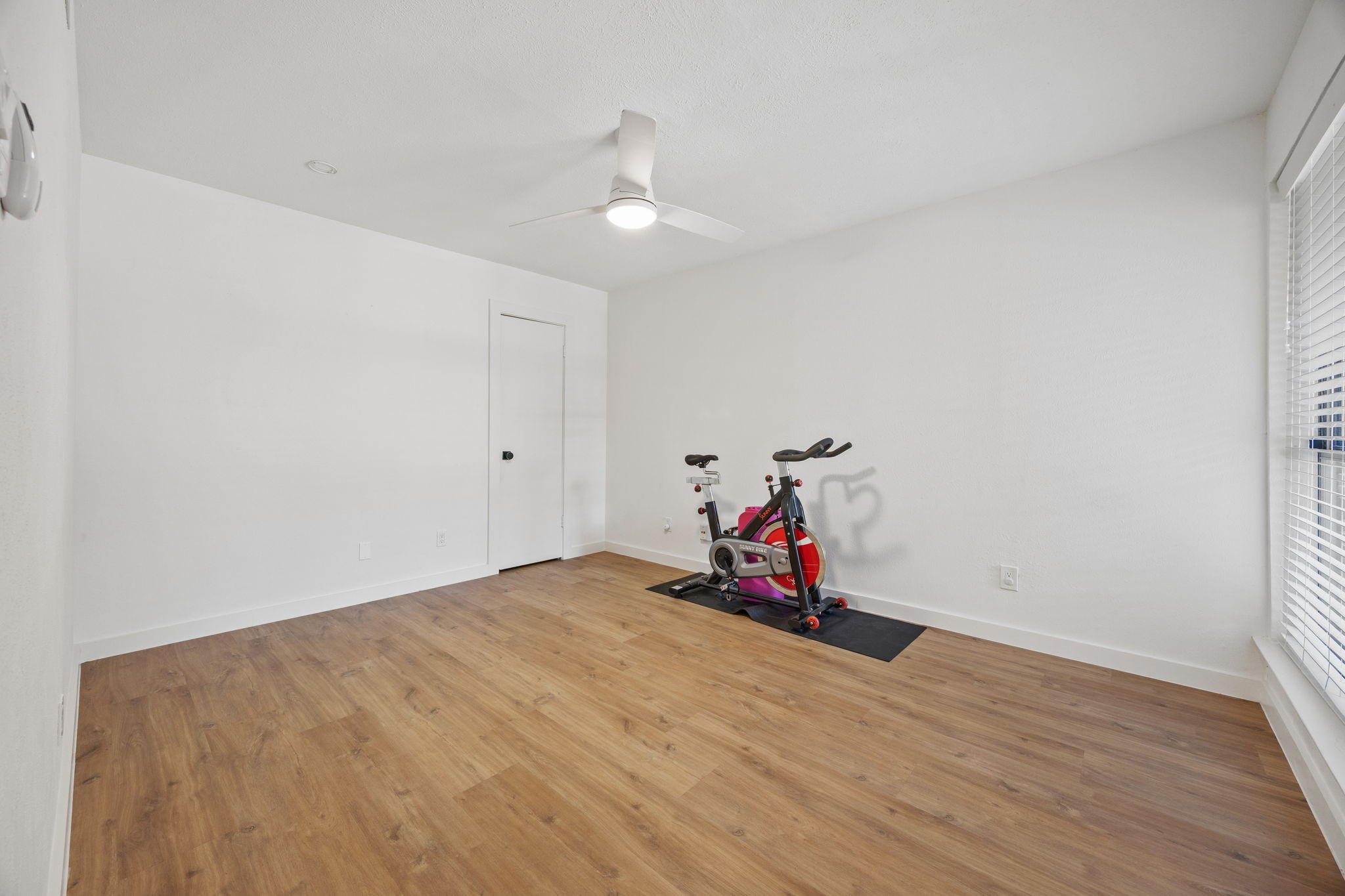 Bright workout room with wood-look flooring, a ceiling fan, and a stationary bike beside a window.