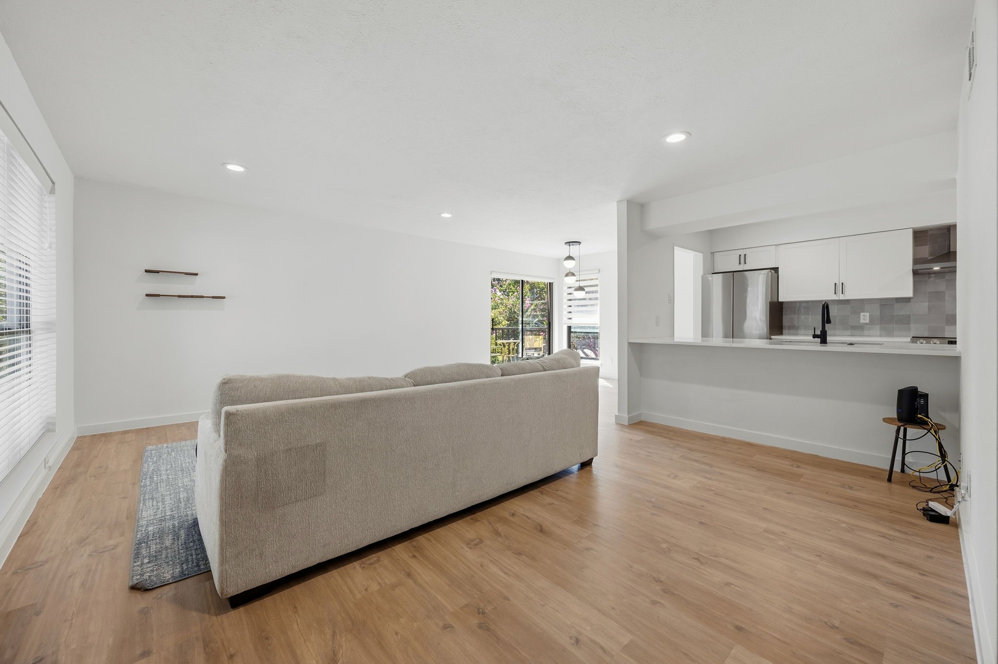 Open-plan living room with a beige sofa, wood floors, and white kitchen with stainless steel appliances.