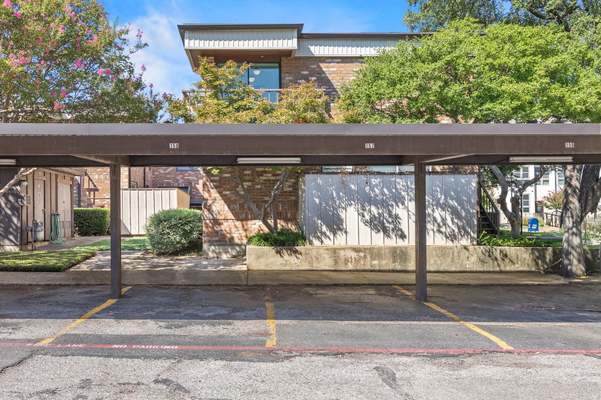 Brick condominium exterior behind covered parking spaces, shaded by mature trees and flowering shrubs.