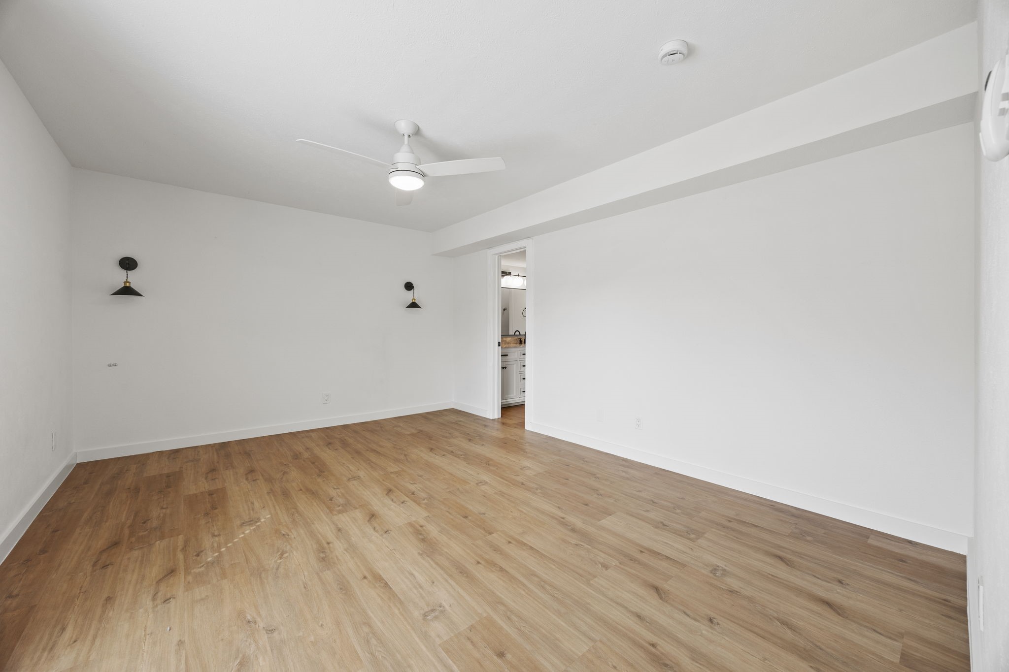 Empty bedroom with light wood floors, black wall sconces, and an open doorway to the bathroom.