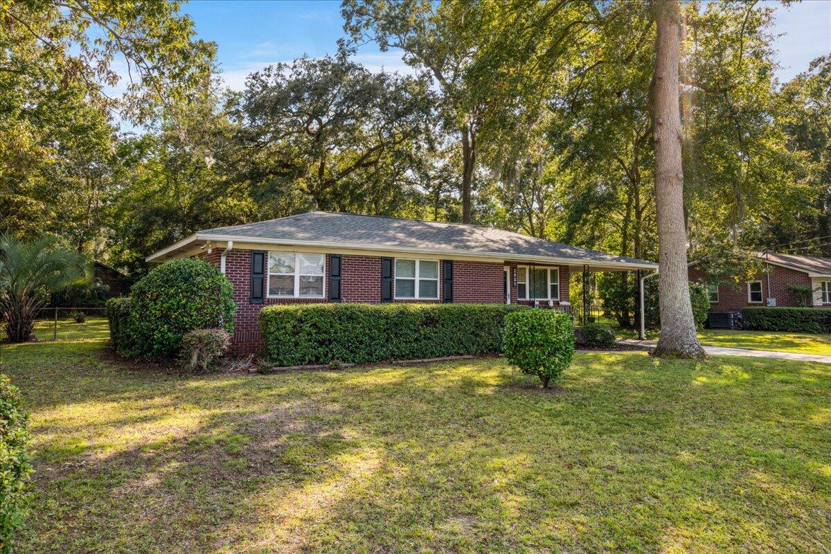 Single-story brick ranch with dark shutters, front hedges, and a covered carport beneath mature trees.