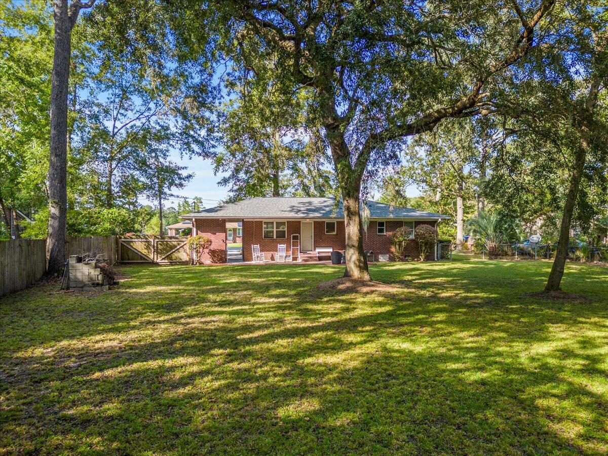 Brick ranch house beyond a shady backyard, with mature trees, a fenced gate, and patio seating.