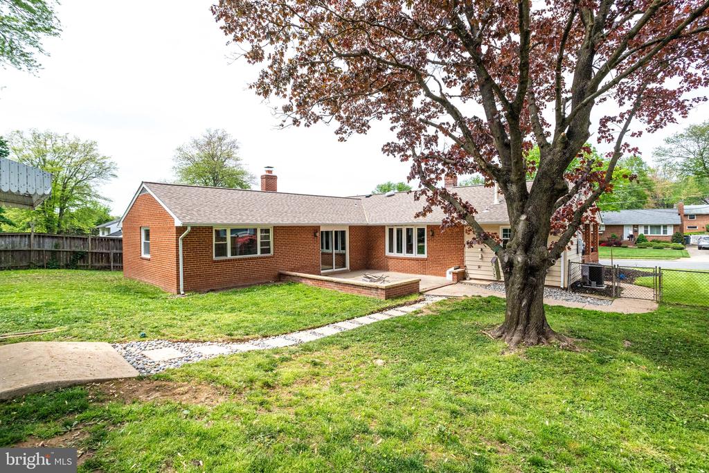 Rear of a single-story brick home with a raised patio, broad lawn, and mature tree in the foreground.