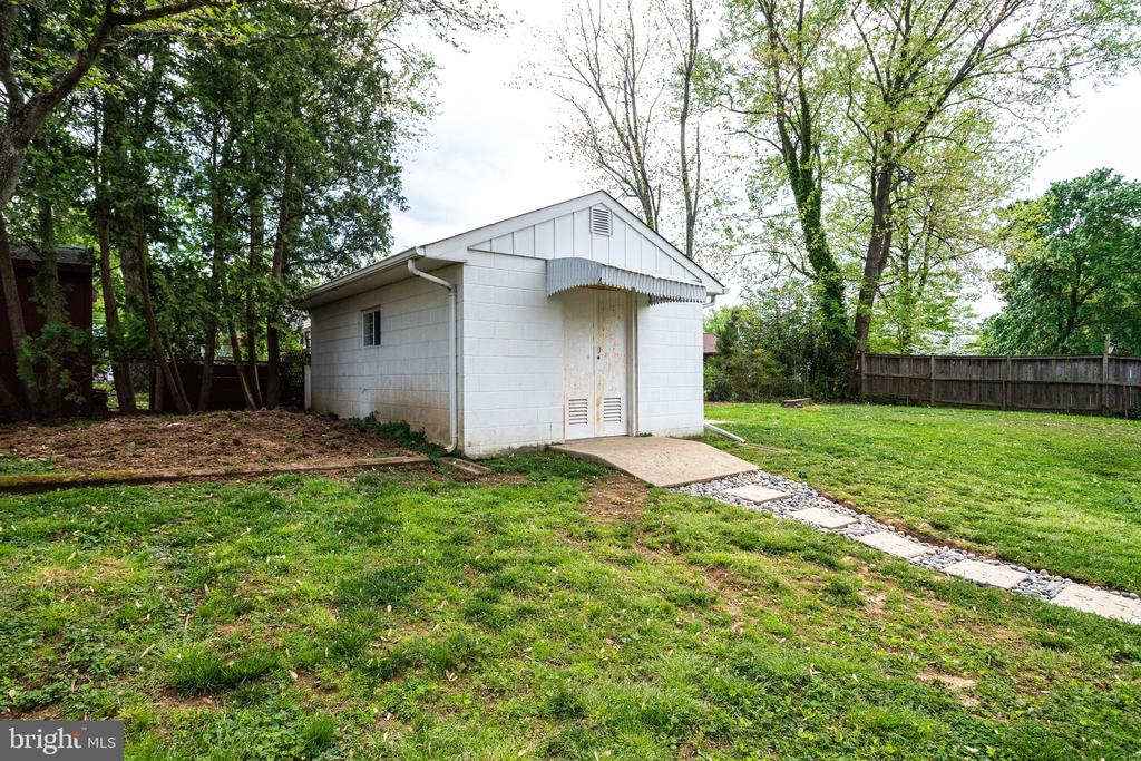 White backyard shed with a gabled roof, stone path, mature trees, and wooden fence.