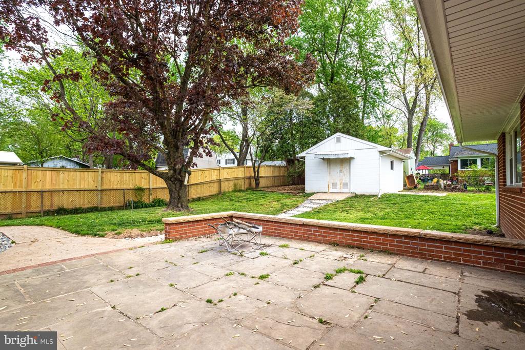 Backyard patio with a brick retaining wall, fenced lawn, mature tree, and detached white shed.