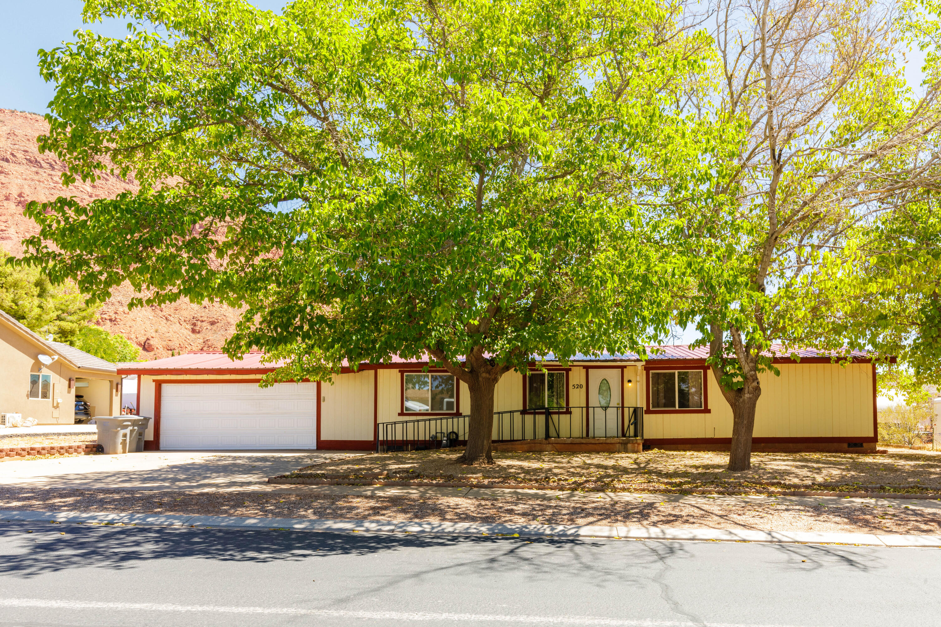 Single-story yellow home with red trim and garage, shaded by large trees beneath a red-rock hillside.