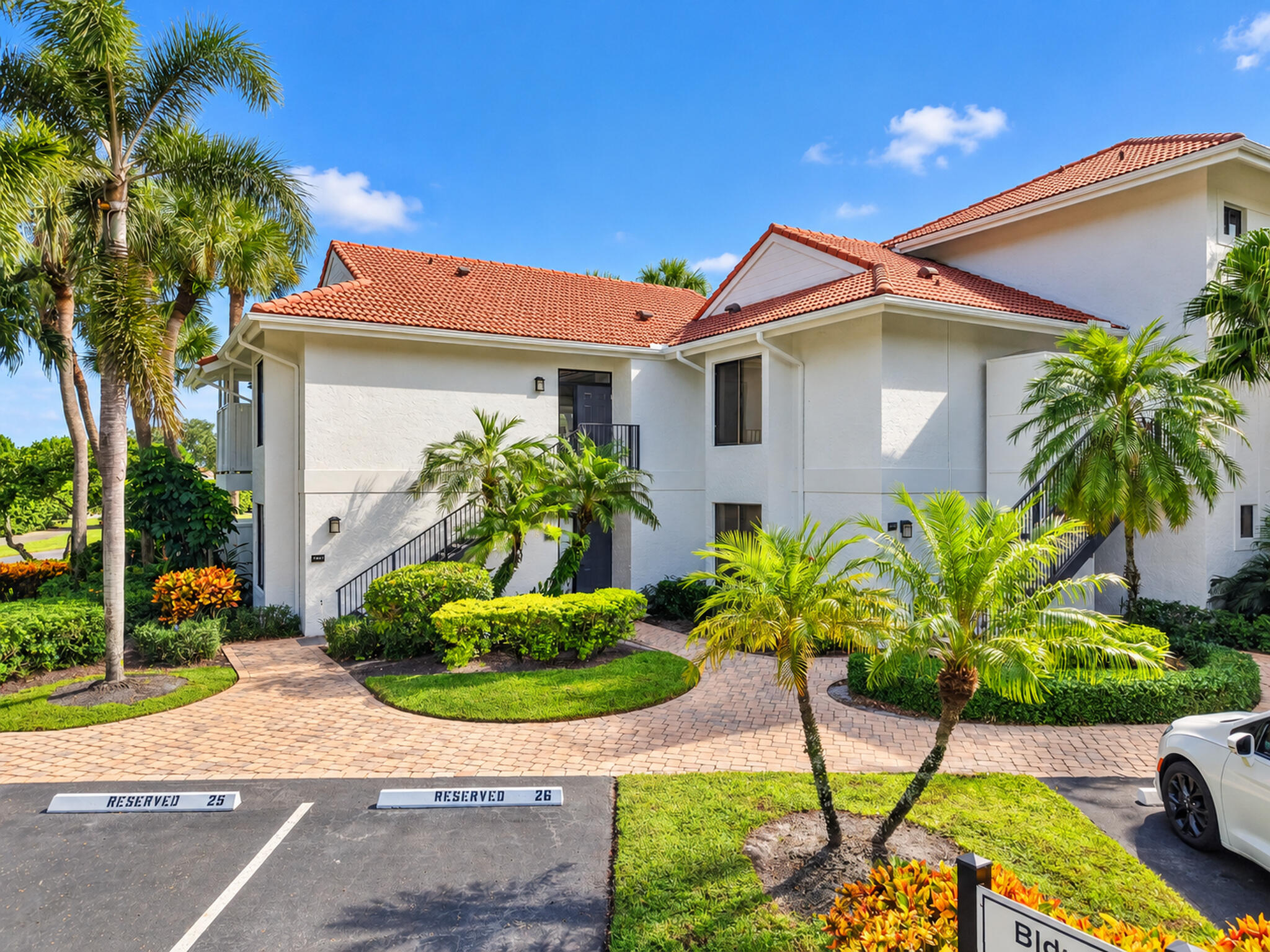 White stucco condominium building with red tile roofs, landscaped palms, and reserved parking spaces.