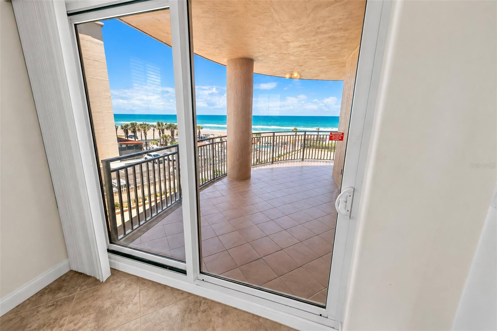 Covered tiled balcony overlooking the beach and ocean, viewed through sliding glass doors.