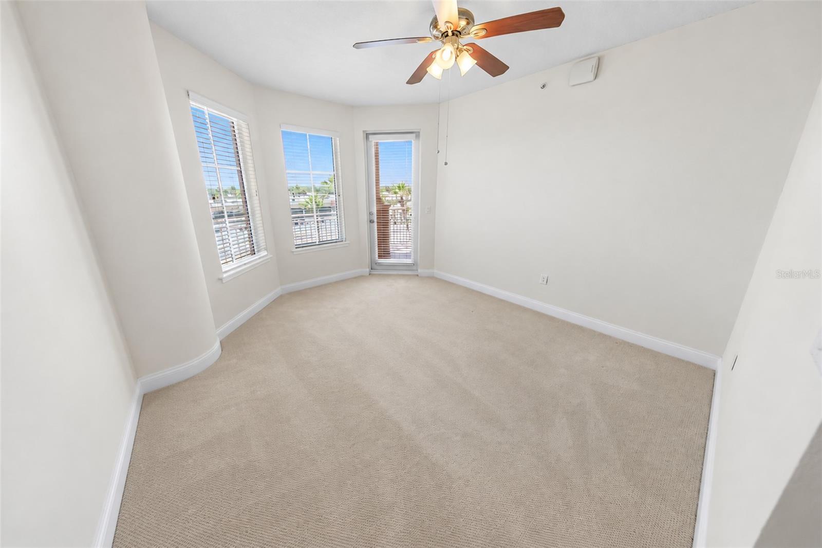 Empty carpeted room with a ceiling fan and windows opening toward a balcony.