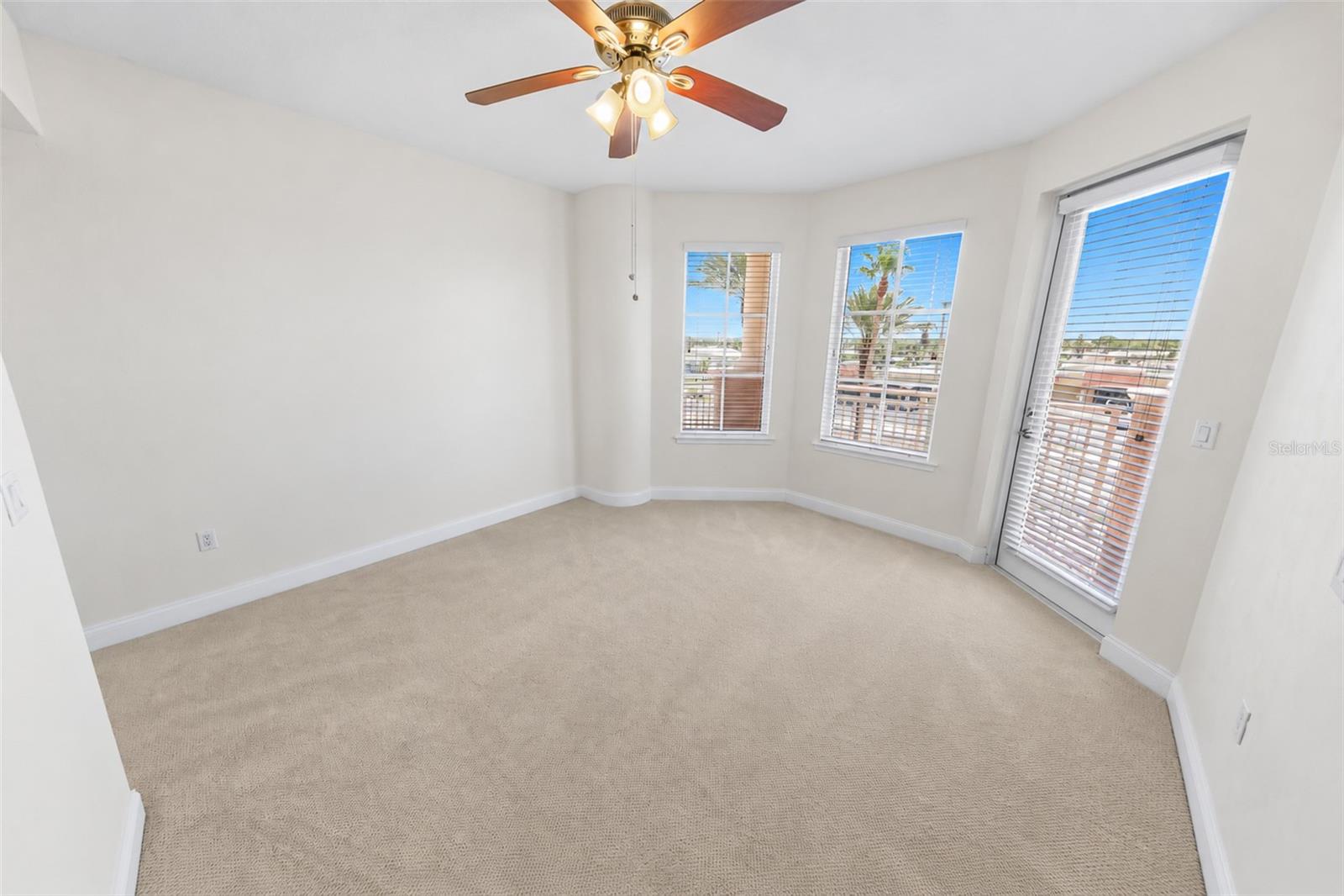 Empty carpeted room with a ceiling fan and windows beside a glass balcony door.