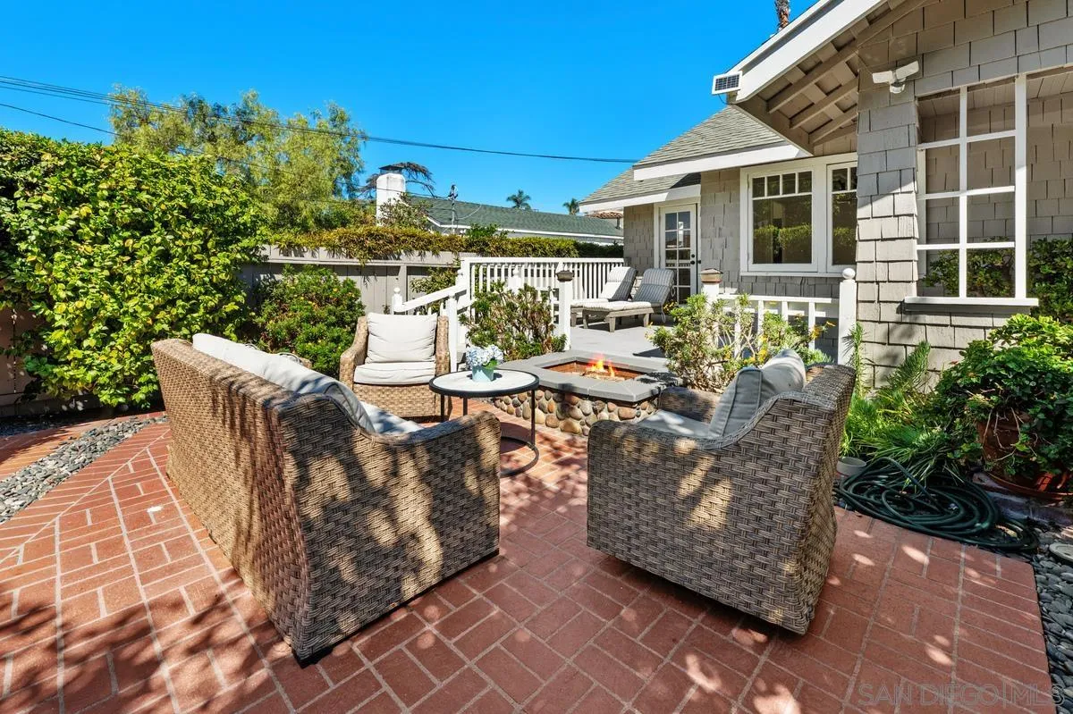 Brick patio with wicker seating and a built-in fire feature beside a gray shingle-style home.