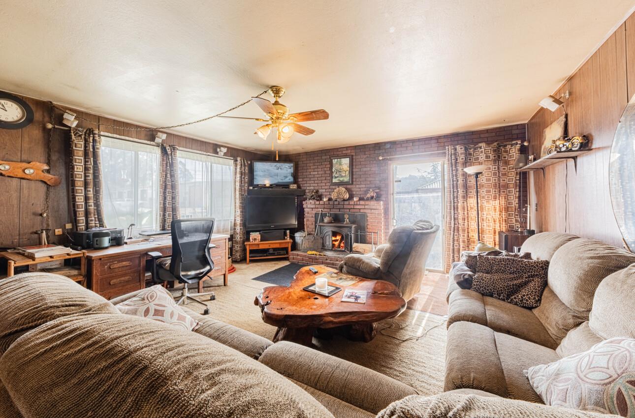 Living room with a brick fireplace, wood-paneled walls, large windows, and a rustic coffee table.