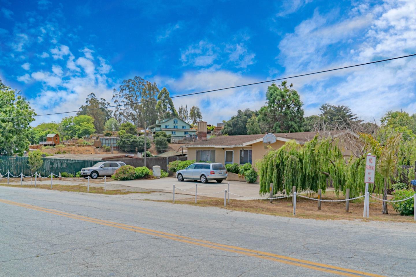 Single-story beige home with a driveway, two parked vehicles, and a drooping evergreen by the road.