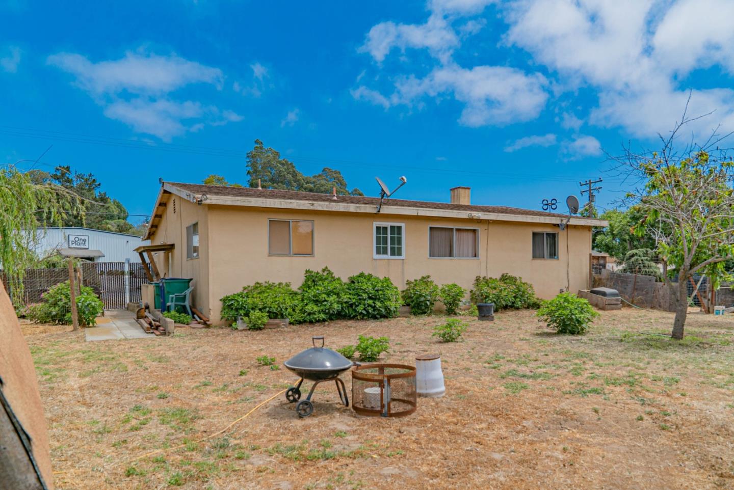Single-story beige house behind a dry backyard with shrubs, a fire pit, and a barbecue grill.