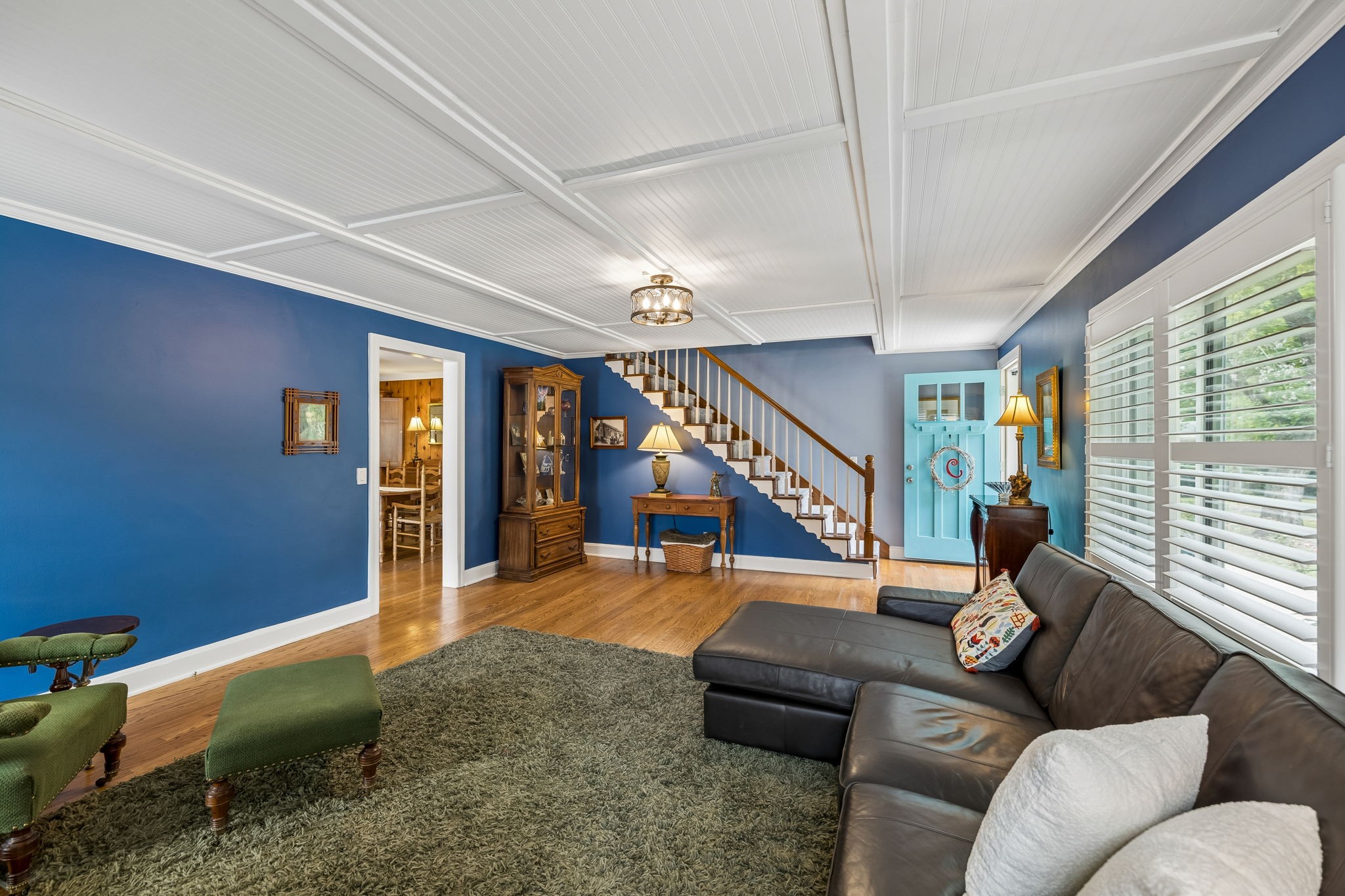 Furnished living room with blue walls, a staircase, and large windows with white shutters.