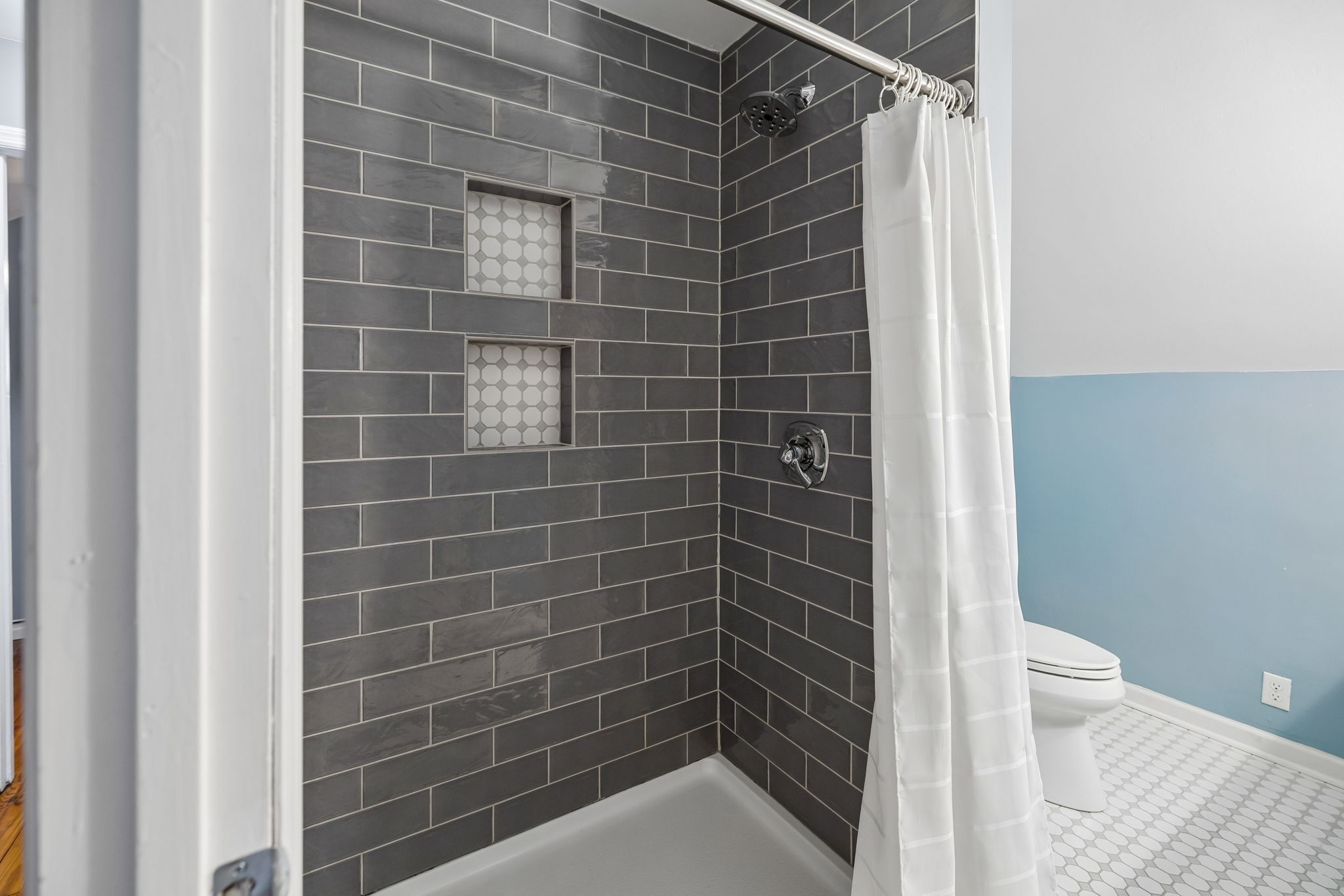 Bathroom with a dark tiled shower, two hexagon-tile niches, and a light blue wall beside the toilet.