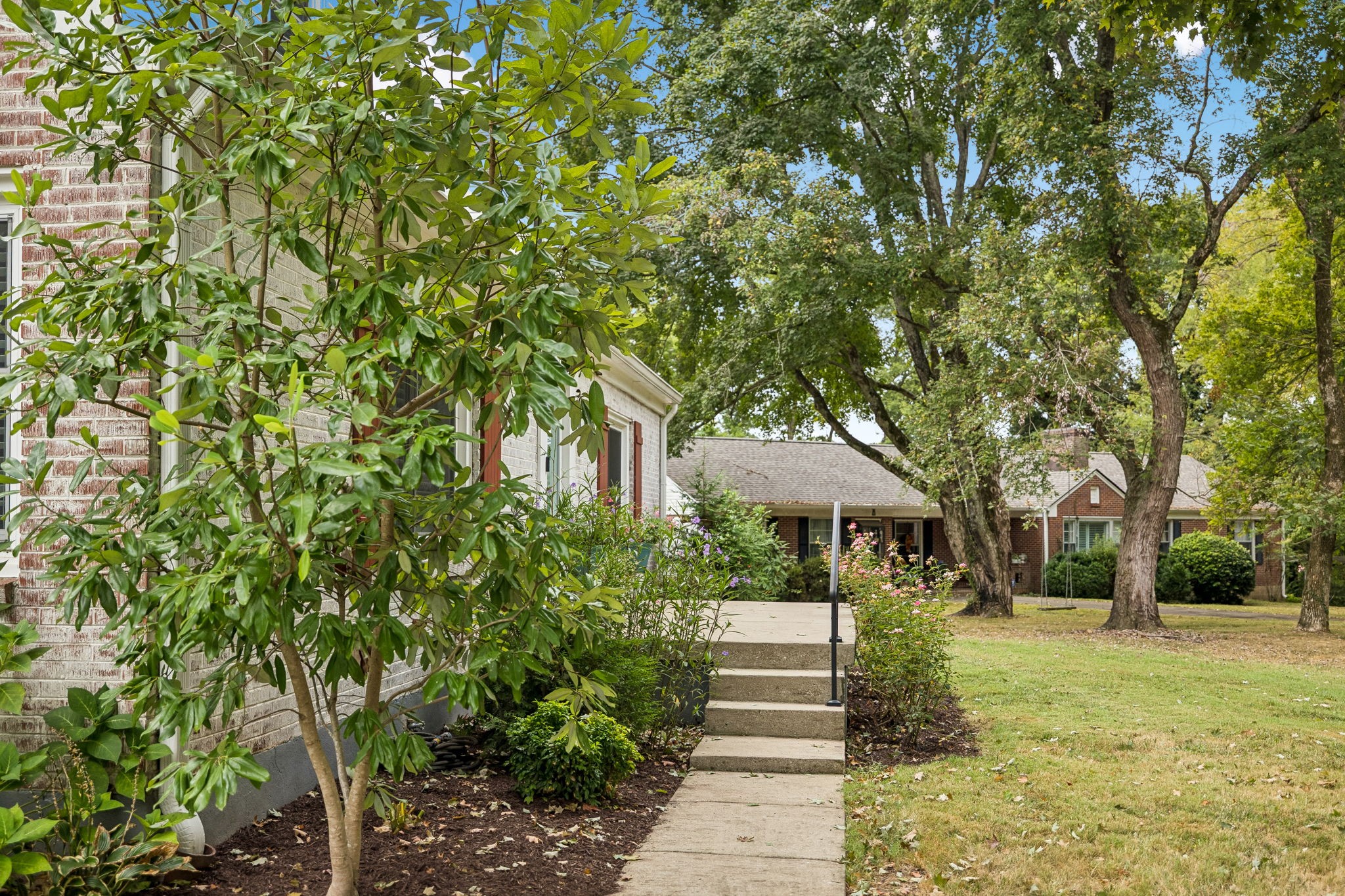 Brick home beside a landscaped walkway, with flowering shrubs and mature shade trees.