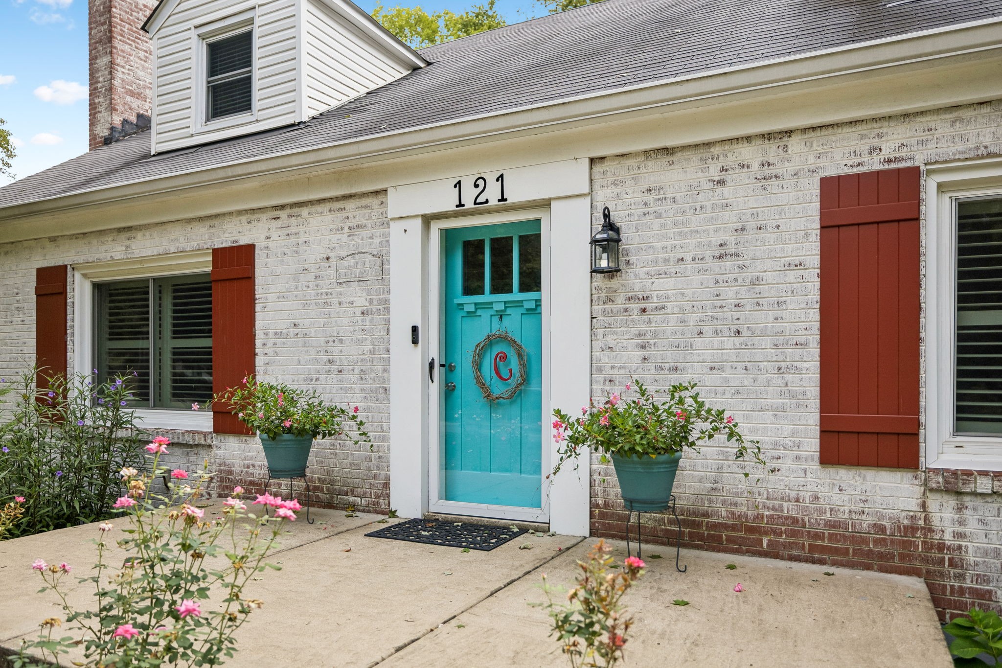 House entrance with a turquoise door, red shutters, and flowering pots beside the walkway.