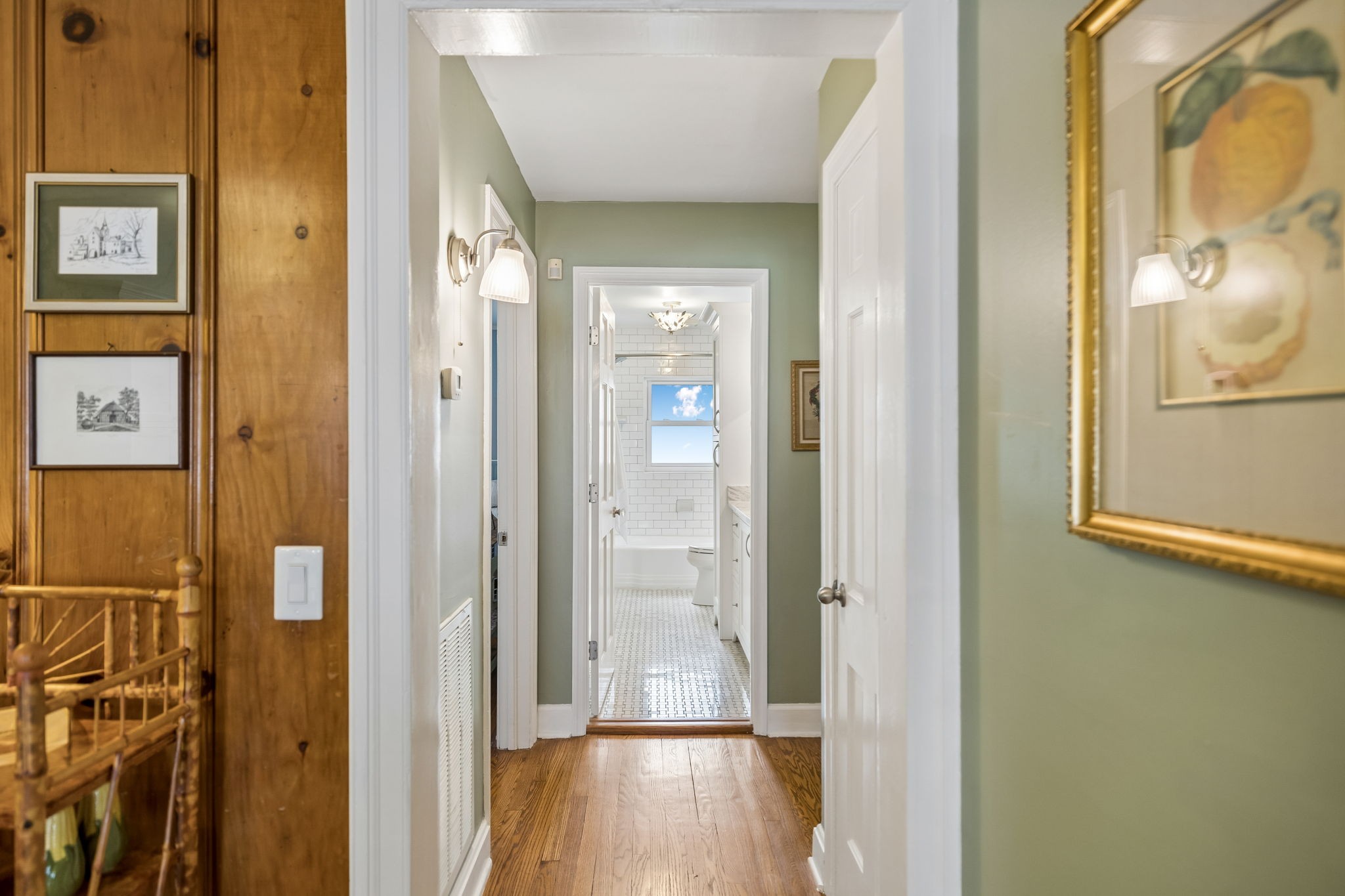 Hallway with hardwood floors and sage-green walls leading to a bright, white-tiled bathroom.