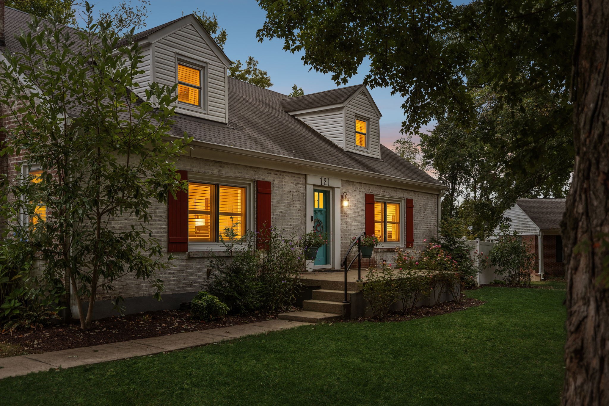 Brick house at dusk with white dormers, red shutters, and a teal front door.