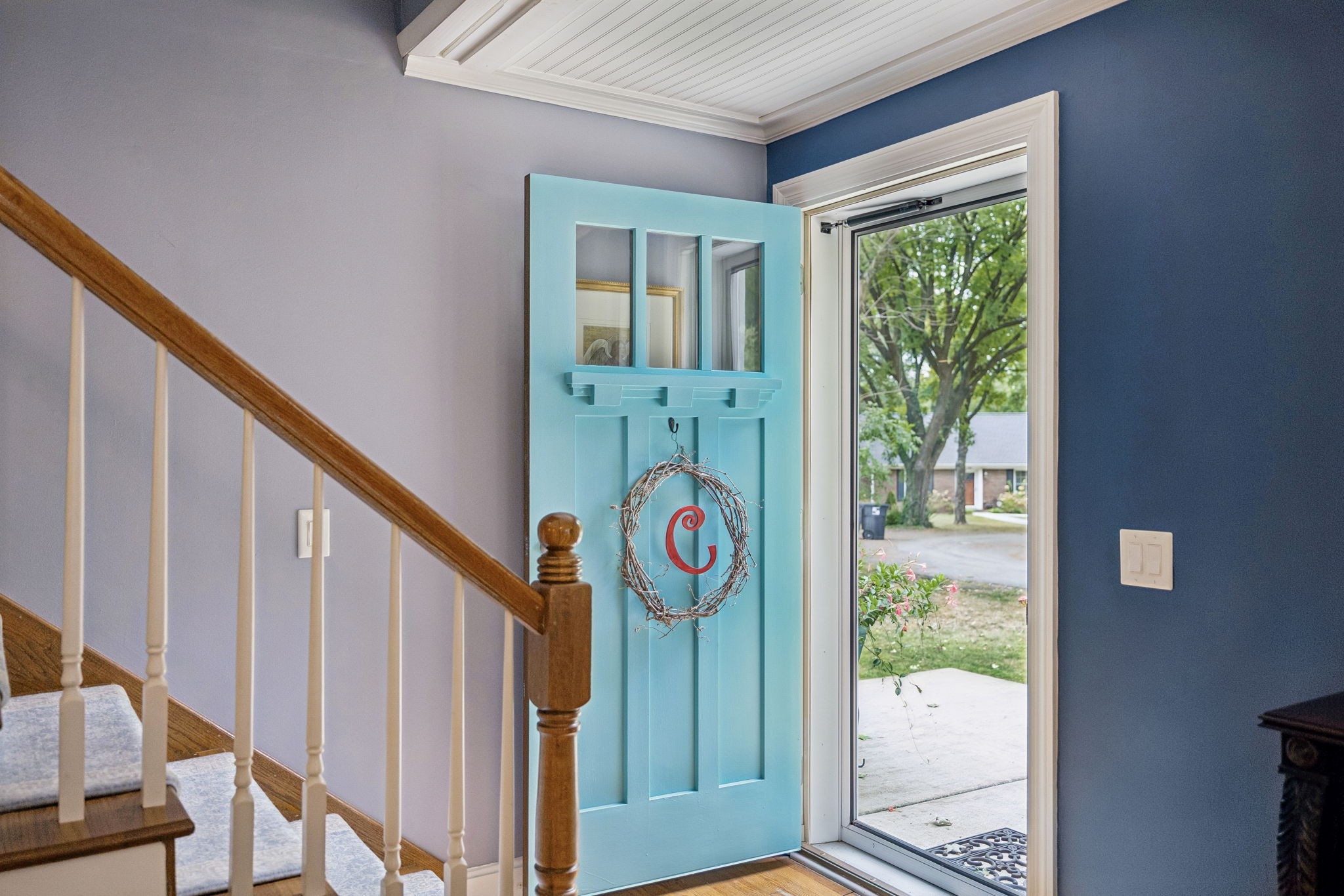 Entryway with an open turquoise front door bearing a wreath beside a wood-and-white staircase.