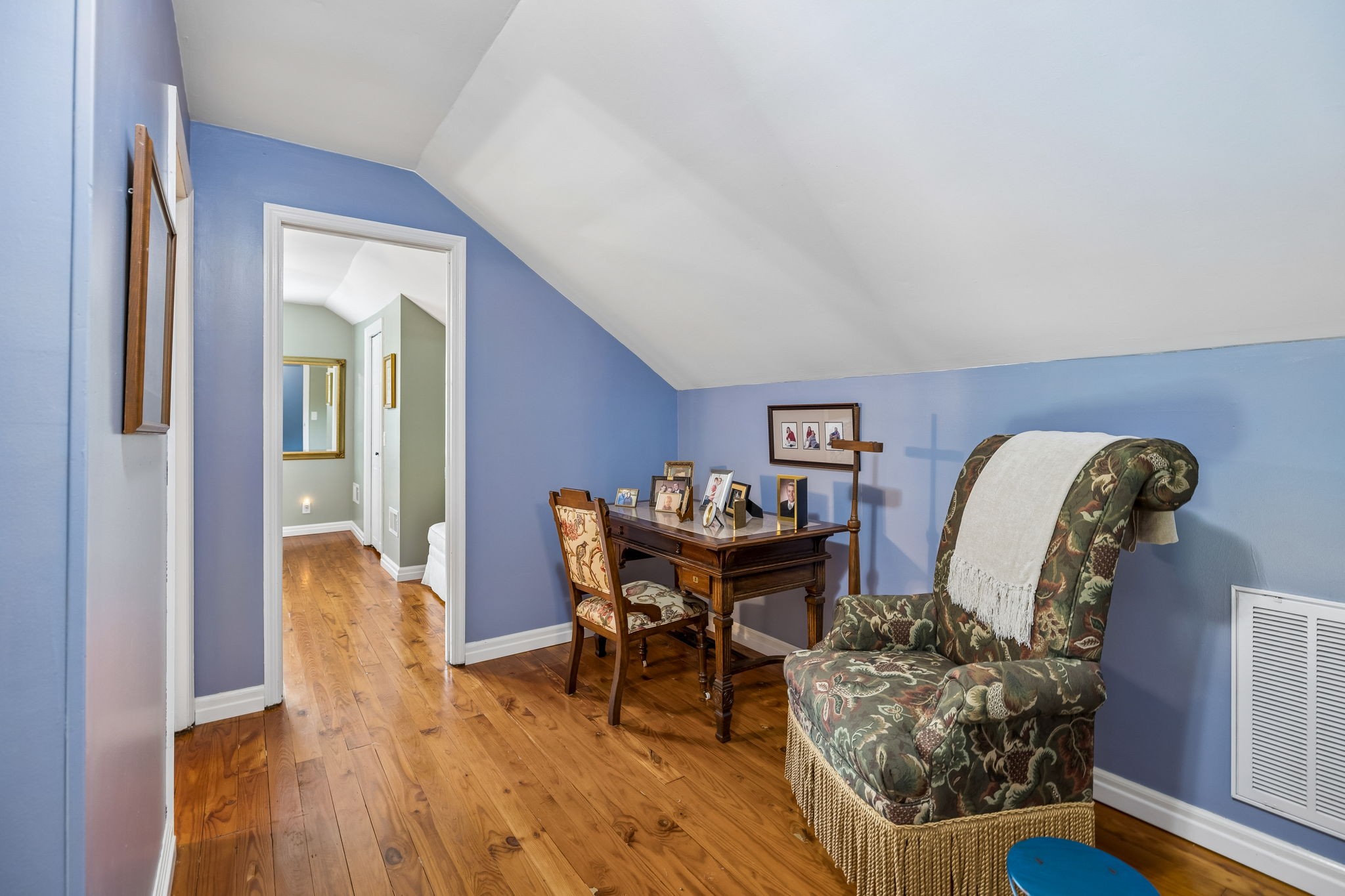 Attic sitting area with a wooden desk, patterned armchair, blue walls, and hardwood floors.