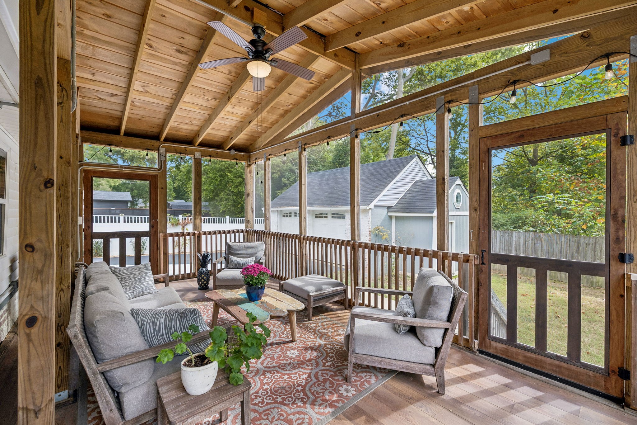 Screened porch with a wood ceiling, gray seating, string lights, and views of trees and a fenced yard.