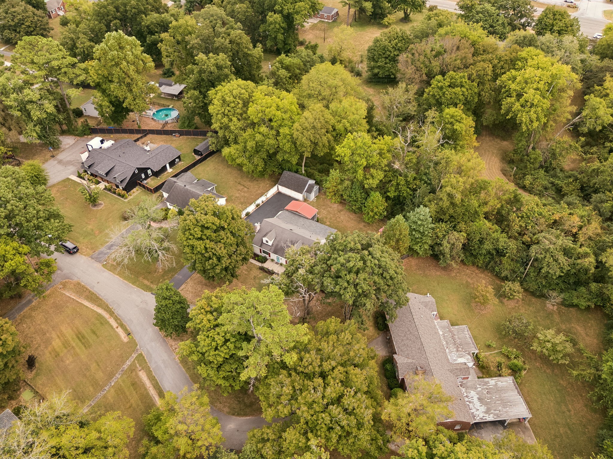 Aerial view of a dark-roofed home among mature trees, with a fenced yard and above-ground pool.