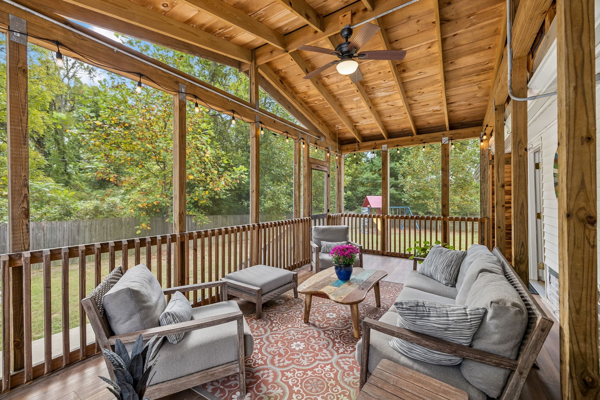Screened-in porch with a wood ceiling, gray seating, string lights, and views of leafy trees.