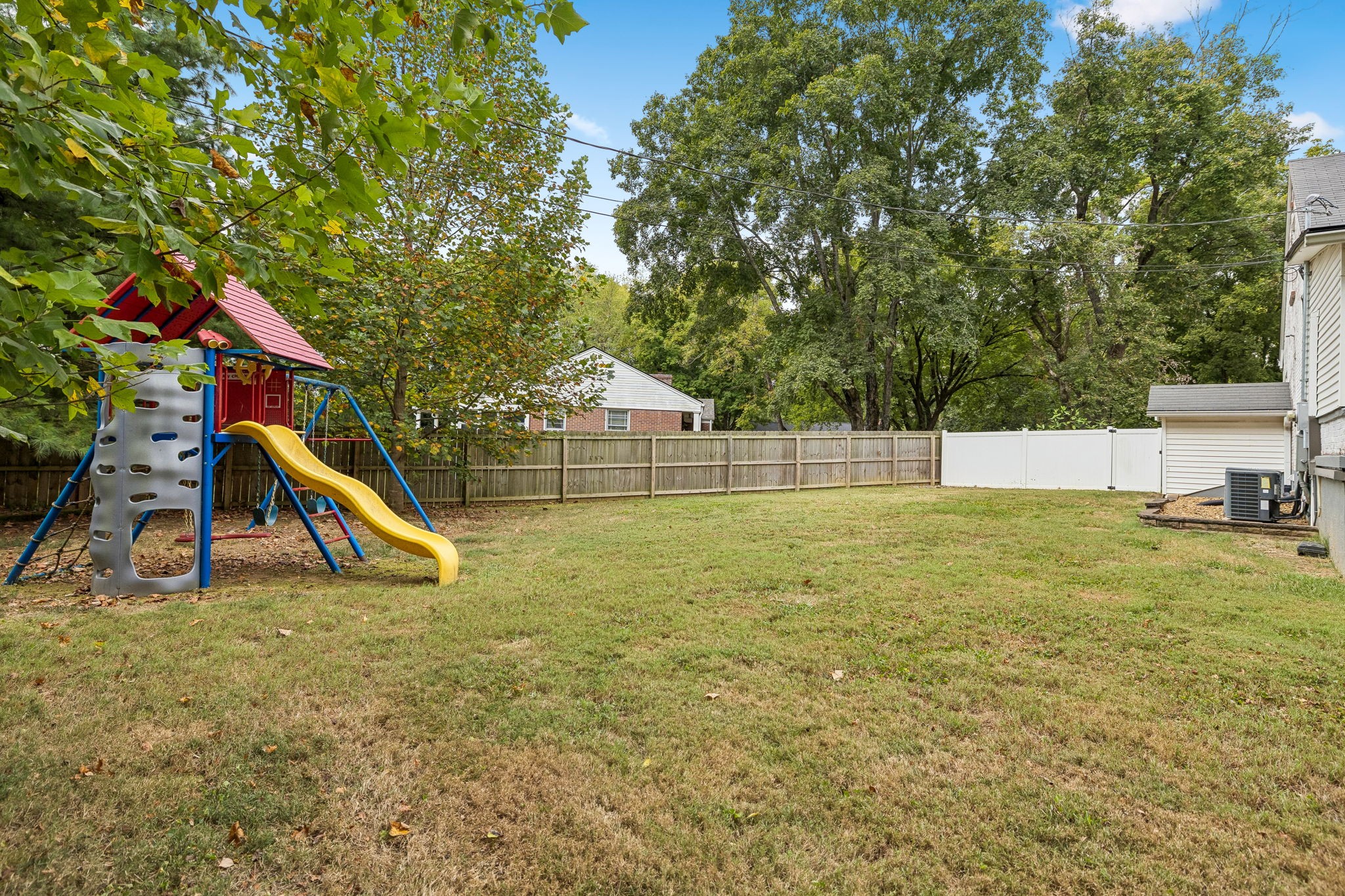 Fenced backyard with a colorful playset and yellow slide beneath mature trees.
