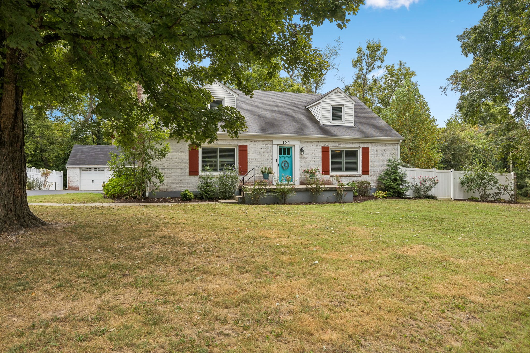 White brick house with turquoise front door, red shutters, dormer, detached garage, and white fence.
