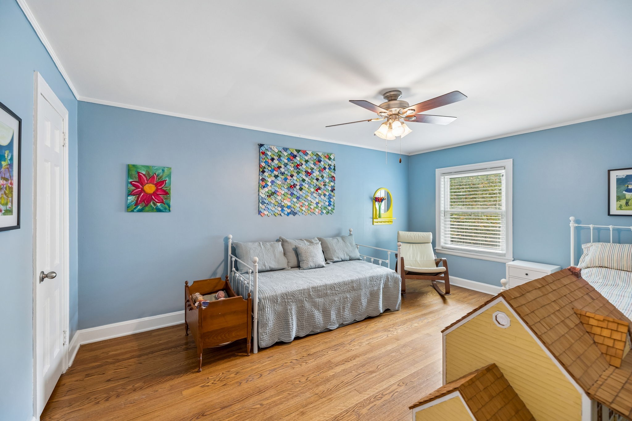 Blue bedroom with hardwood floors, a white daybed, ceiling fan, and a dollhouse in the foreground.