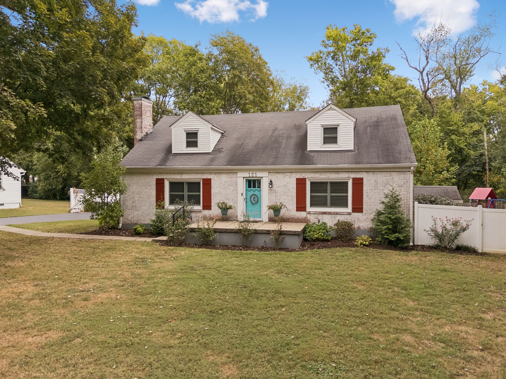 White brick house with red shutters and a turquoise door, framed by mature trees and a broad lawn.