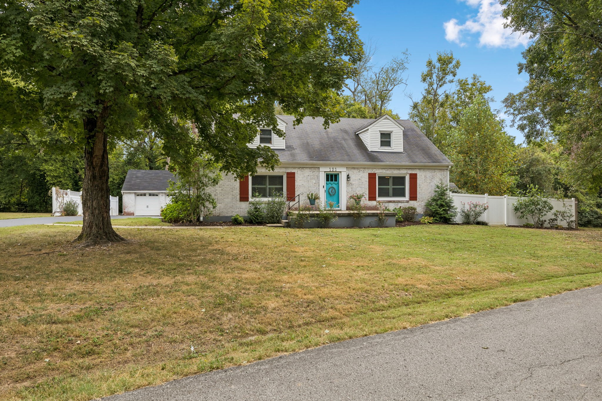 White brick house with a turquoise front door, red shutters, dormers, detached garage, and fenced yard.