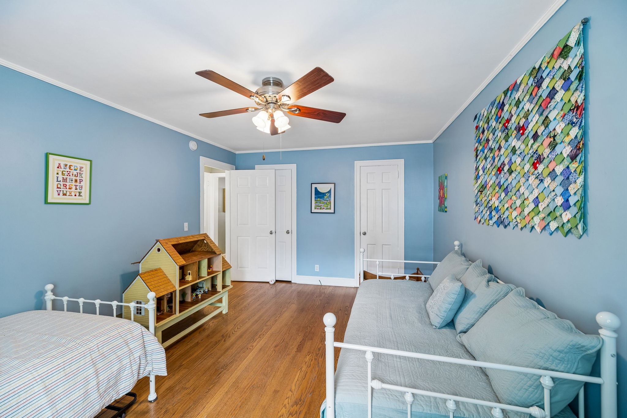 Blue bedroom with two white beds, a wooden dollhouse, and colorful fabric wall art.