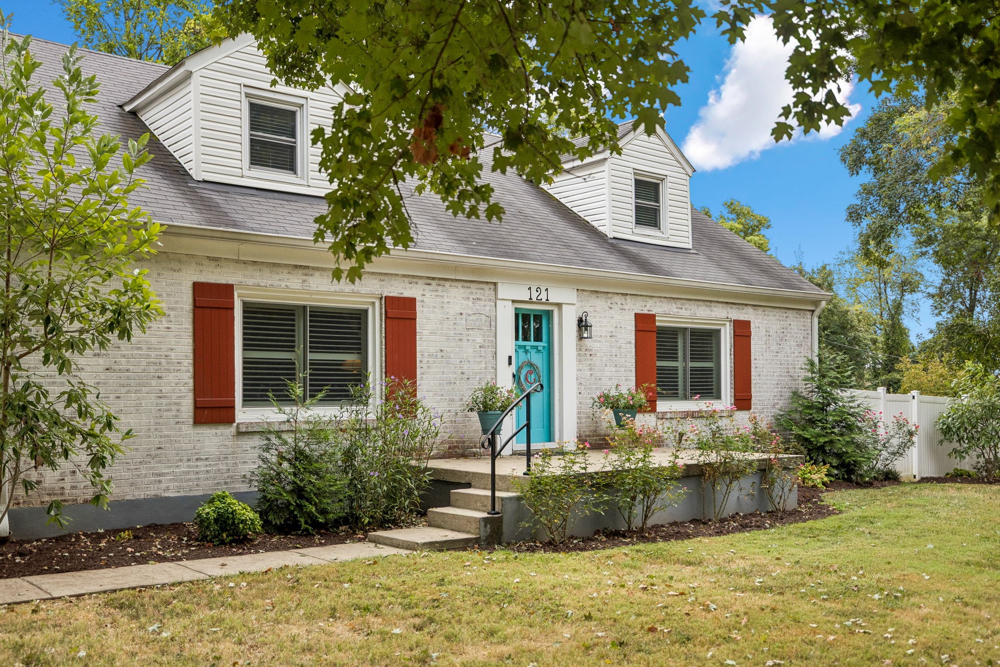 Light brick house with red shutters, dormers, and a turquoise front door framed by leafy trees.