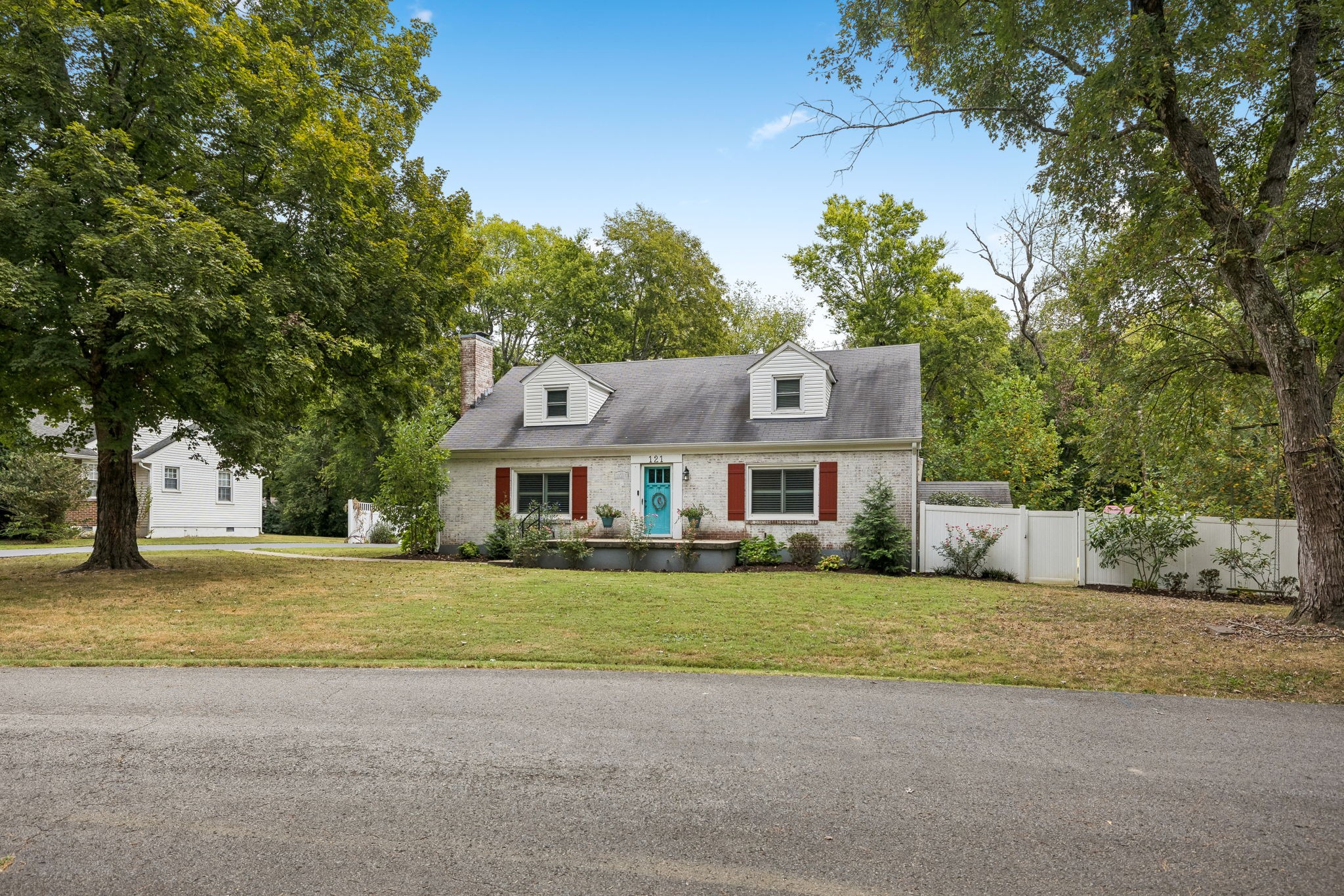 White-brick home with turquoise front door, dormers, mature trees, and a white yard fence.