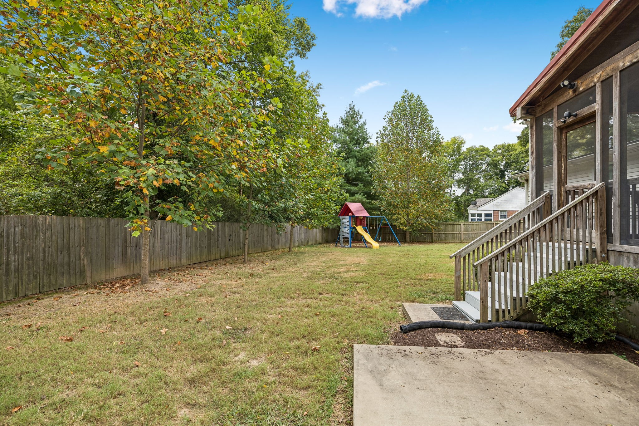 Fenced backyard with mature trees, a children's playset, and wooden deck stairs.