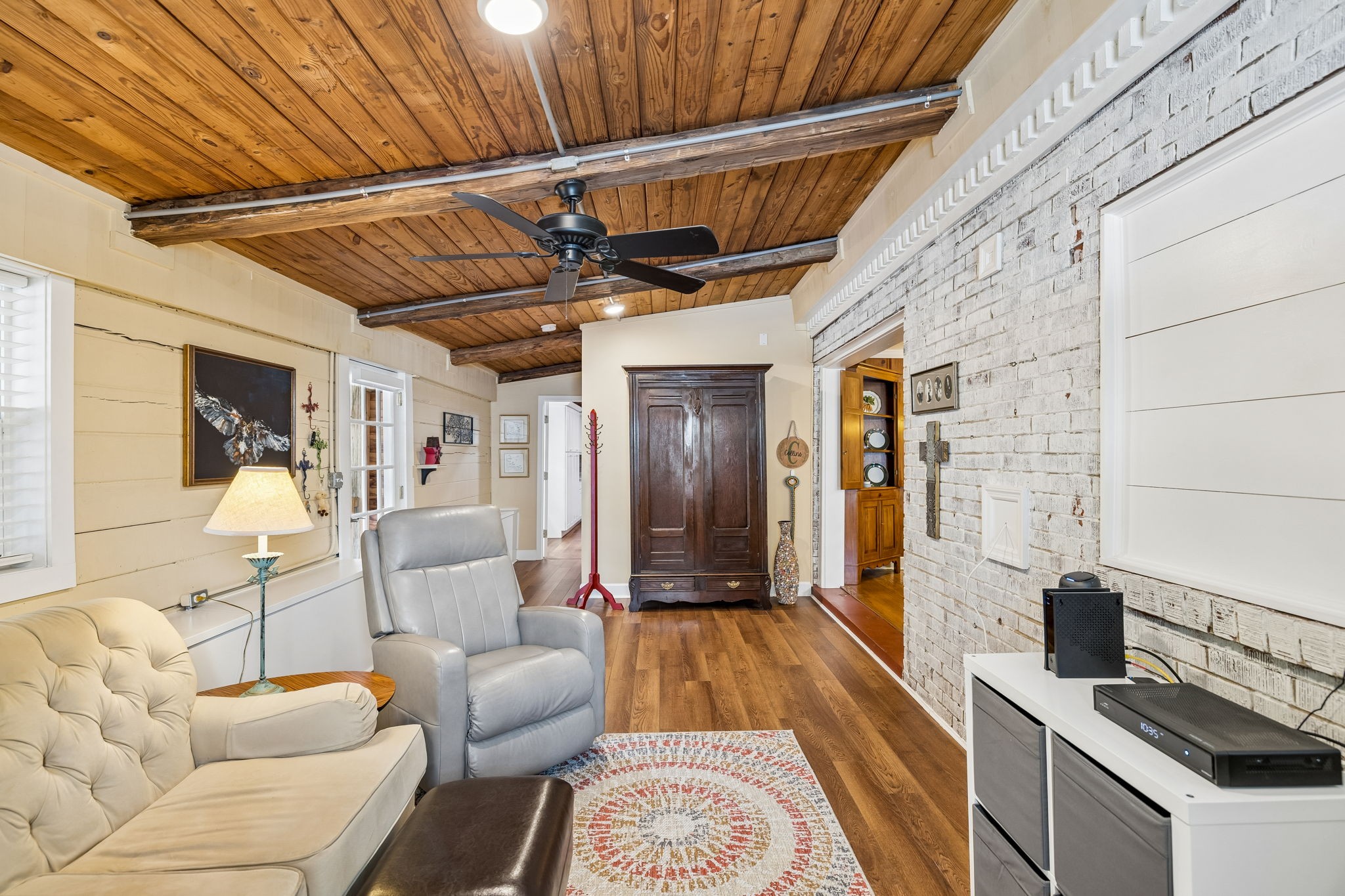 Living room with a vaulted wood ceiling, exposed beams, whitewashed brick wall, and hardwood floors.