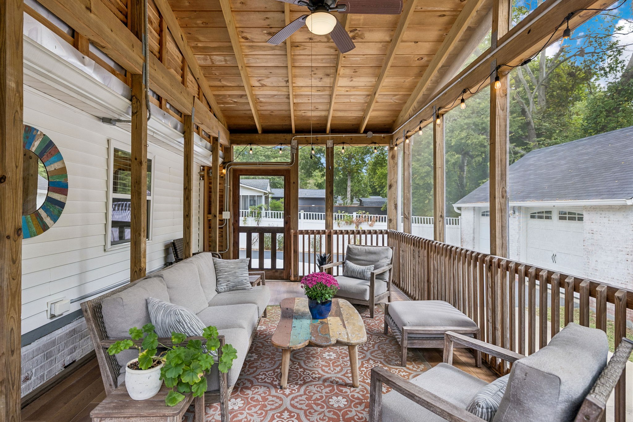 Screened porch with exposed timber ceiling, string lights, and gray outdoor seating around a colorful coffee table.