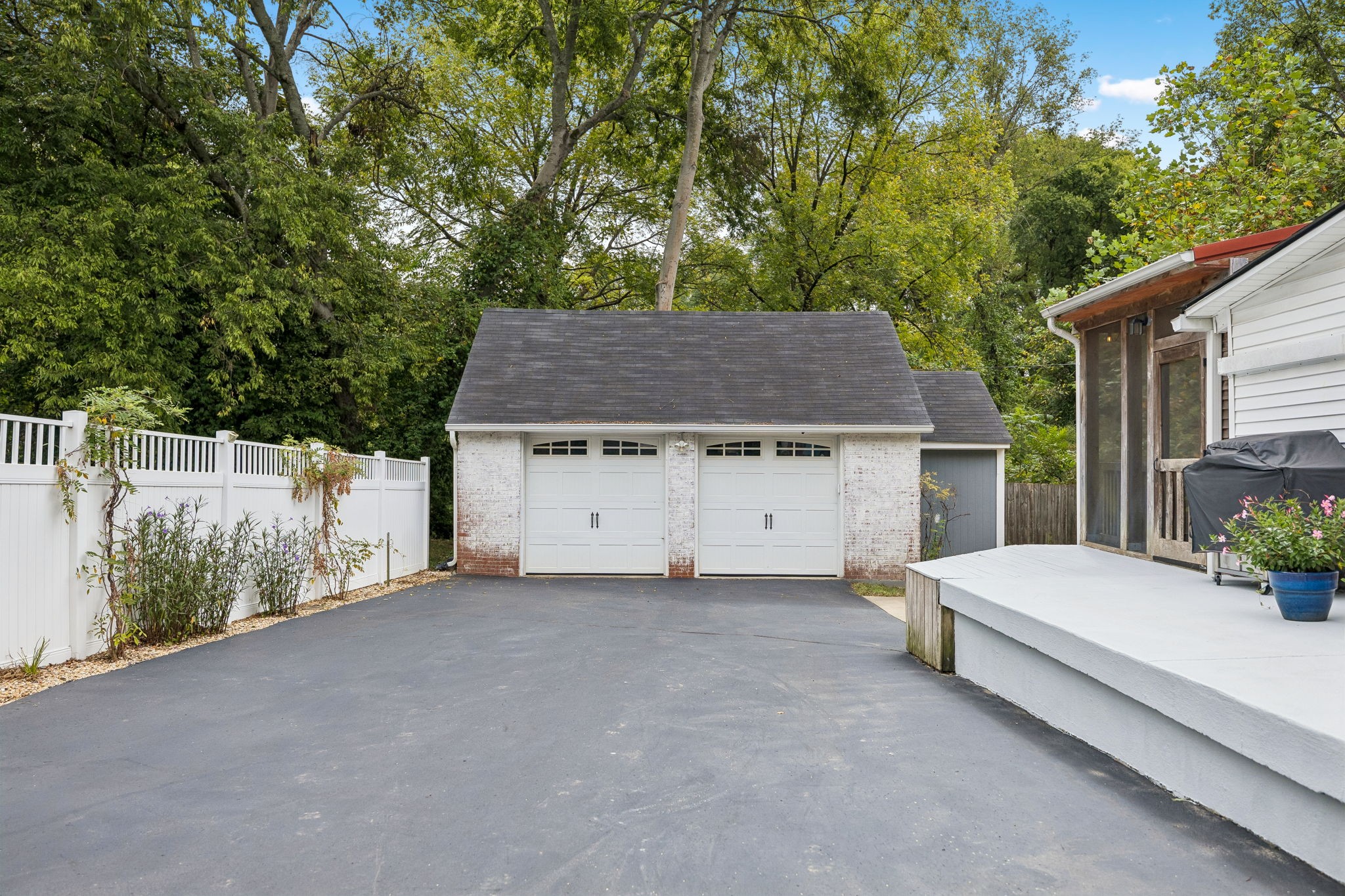 Detached two-car garage at the end of a paved driveway, bordered by a white fence and wooded greenery.